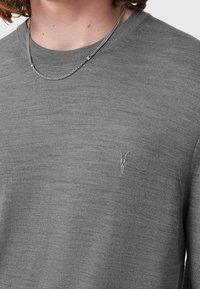 Person, der einen grauen Pullover mit einem kleinen gestickten Logo eines gehörnten Tieres und einer silbernen Kette trägt.