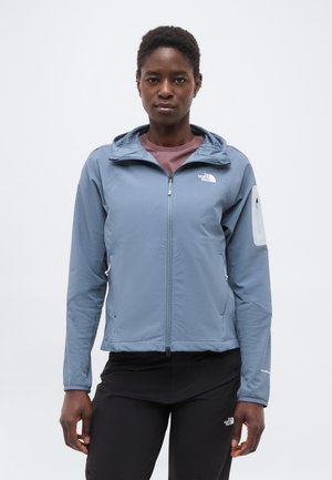 Persoană purtând o jachetă albastră cu fermoar și glugă și pantaloni negri, stând în picioare în fața unui fundal simplu, deschis la culoare, privirea îndreptată înainte.