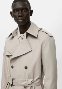 Beżowy trencz z dwurzędowym zapięciem, dużym kołnierzem, pagonami na ramionach oraz pasem w tym samym kolorze. Gładka faktura materiału i guziki.