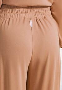 Pantalones beige con cintura elástica y tela suave y elástica. Cuenta con una pequeña etiqueta de marca rosa en la cintura. Diseño sencillo con detalles mínimos.