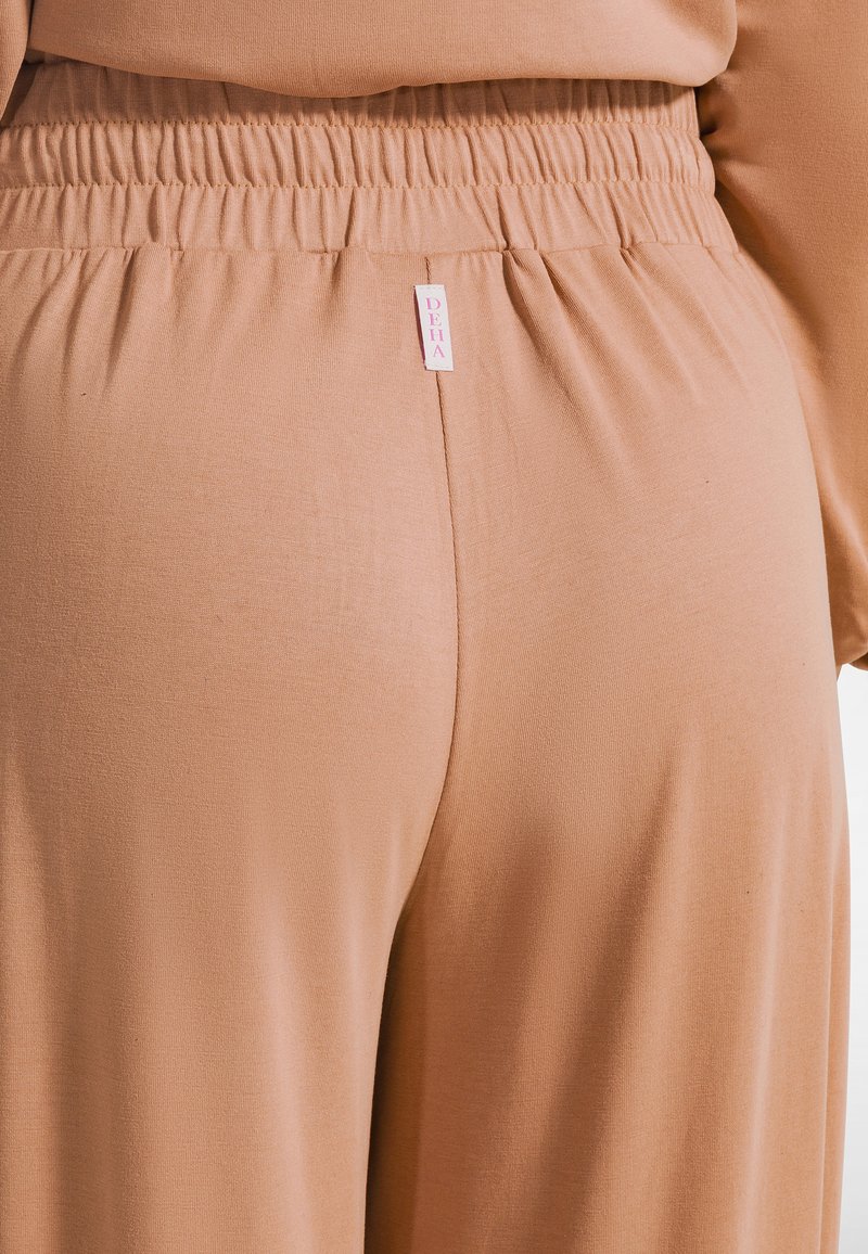 Pantalones beige con cintura elástica y tela suave y elástica. Cuenta con una pequeña etiqueta de marca rosa en la cintura. Diseño sencillo con detalles mínimos.