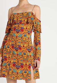 Vestido sin hombros de vibrante tela naranja, con patrones florales en rojo y verde, detalles con volantes y aberturas laterales.