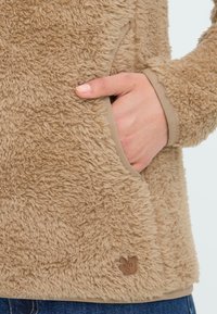 Beige Fleecejacke mit einer weichen, strukturierten Oberfläche, an der eine Hand in einer seitlichen Tasche steckt und einem kleinen braunen Logotag am Saum.