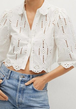 Blouse blanche courte avec bords festonnés et broderie anglaise portée avec un jean taille haute bleu clair, mains dans les poches.