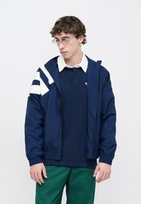 Temno modra vetrovka z belo črtasto obrobo, z zadrgo in elastičnimi manšetami, nošena čez temno modro polo majico in zelene hlače. Prozorna sončna očala.