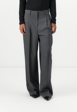 Pantalons gris ajustés avec des plis, fabriqués en tissu lisse. Dotés d'une taille haute et d'un design à jambes larges. Associés à des chaussures noires à bout pointu.