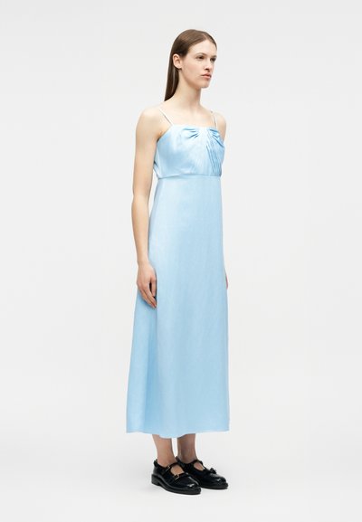 Νεαρή γυναίκα όρθια, φοράει αμάνικο ανοιχτό μπλε midi φόρεμα με πλισέ μπούστο και μαύρα παπούτσια Mary Jane, απλό λευκό φόντο.