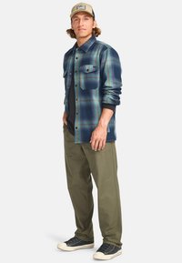Ung man som bär en beige keps, en blå och grön rutiga skjorta över en svart topp, olivgröna byxor och svarta sneakers, stående mot en enfärgad bakgrund.