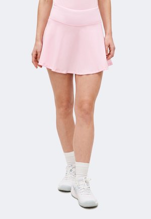 Žena v svetloružovej tenisovej sukni, bielych ponožkách a bielych športových topánkach, stojaca so skríženými nohami na bielom pozadí.