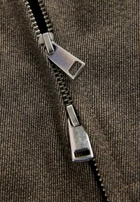 Tessuto marrone con una cerniera dotata di due tiranti metallici. I denti della cerniera sono visibili lungo il bordo, mostrando una finitura durevole e testurizzata.