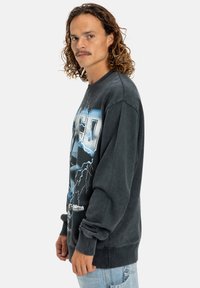 Sudadera gráfica negra con un patrón de rayos, que presenta un diseño impreso y puños acanalados, con un corte holgado, combinada con jeans claros.