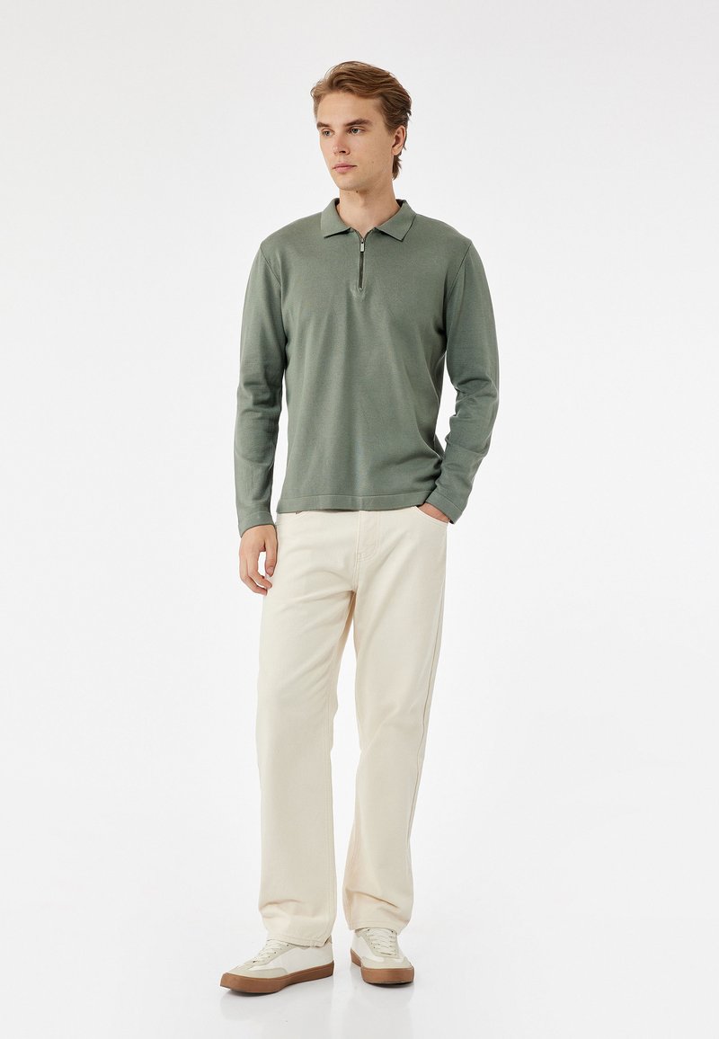 Polo de manga larga verde claro con cuello de media cremallera, combinado con pantalones color crema y zapatillas blancas con suelas marrones.