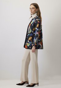 Giacca nera trapuntata con motivi astratti colorati, abbinata a pantaloni larghi crema e décolleté nere a punta.