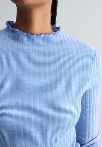 Femme portant un haut à rayures verticales bleues et blanches avec un col à volants, cheveux tressés visibles à l'arrière sur un fond uni.