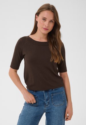 Femme aux longs cheveux bruns portant un haut marron foncé à manches courtes et un jean bleu avec des poches plaquées à l'avant, debout devant un fond uni.