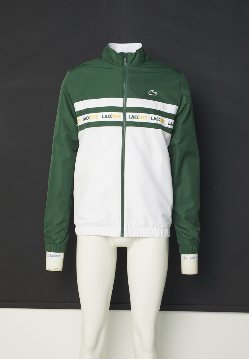 Veste coupe-vent verte et blanche avec une fermeture éclair intégrale, un col montant et des poignets élastiques ; présente un logo Lacoste horizontal sur la poitrine.