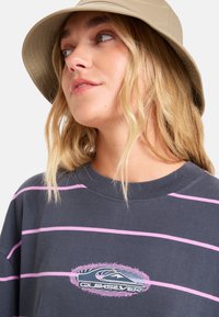 T-shirt gris rayé avec des lignes roses, présentant un logo brodé en forme de cercle. Le modèle porte un chapeau de seau beige, avec des cheveux blonds ondulés visibles.