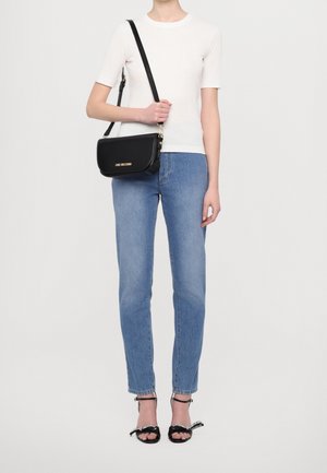 Vrouw in een witte korte mouwen top en blauwe jeans, met een zwarte Love Moschino schoudertas, en zwarte open teen hakken.