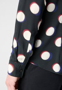 Gros plan d'une chemise noire à manches longues avec de grands cercles blancs bordés de rouge et de bleu, portée avec un pantalon noir.