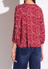Kobieta ubrana w czerwoną, wzorzystą bluzkę na długi rękaw z bufkami oraz niebieskie dżinsy, stojąca tyłem do kamery.