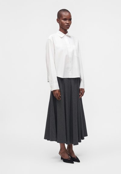 Femme portant une chemise blanche à manches longues boutonnée, une jupe midi plissée gris foncé et des chaussures noires pointues à talon bas, debout sur fond blanc.