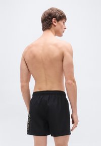 Μαύρα μαγιό σε σορτς με ελαστική μέση και λογότυπο στο πλάι. Ελαφρύ ύφασμα με λεία υφή, εμφανίζεται από πίσω.