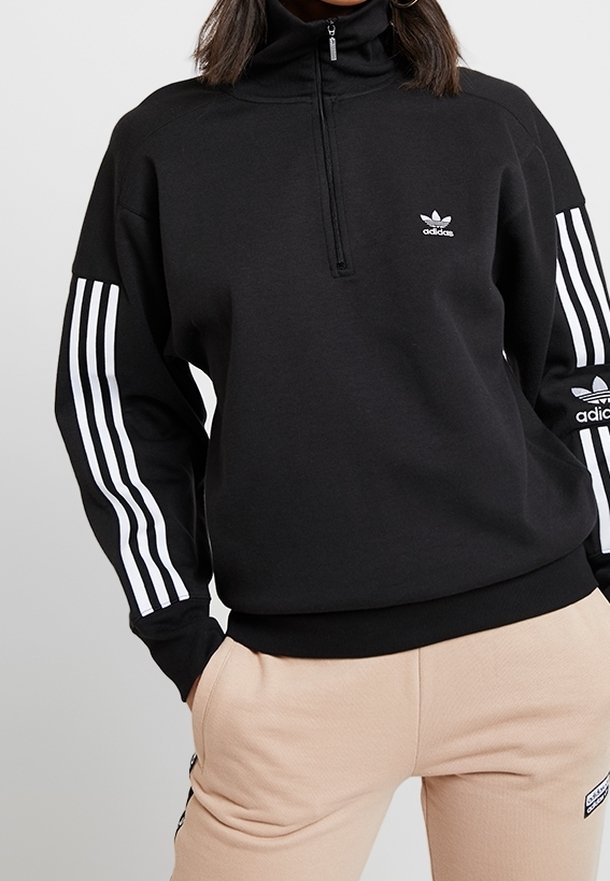 Personne portant un sweat à col zippé noir Adidas avec des bandes blanches sur les manches et un pantalon beige, les mains dans les poches.