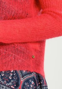 Roter, flauschiger Strickpullover mit lockerem Gewebe, ausgestattet mit einem Akzent aus goldenem Logo. Kombiniert mit einem gemusterten Stoff darunter.