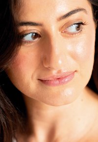 Close-up van een gezicht met een gladde, lichte huid. Ogen zijn bruin, haar is donker, en lippen hebben een lichte glans. De huid vertoont een natuurlijke glans.