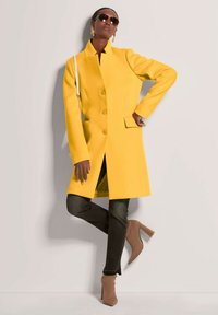 Geel wollen jas met een hoge kraag, knoopsluitingen en zijzakken; gecombineerd met donkere jeans en beige hoge hakken.