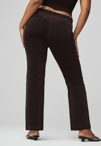 Dunkelbraune Schlagjeans mit Gesäßtaschen, subtilen Nähten und einer glatten Textur. Tragen mit schwarzen Absatzsandalen.