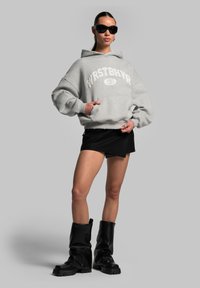 Hoodie overdimensionato grigio con testo "WRSTBHVR", mini gonna nera e stivali alti fino al ginocchio neri. Modello con occhiali da sole neri.