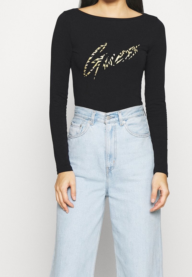 Långärmad svart topp med guldig leopardtrycklogo vid bröstet, i kombination med högmidjade ljusblå jeans. Material i bomullsblandning.