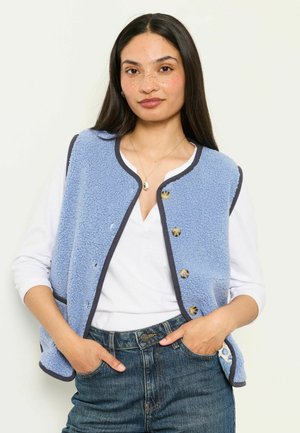 Kvinde med langt mørkt hår iført en lyseblå fleecervest med knapper over en hvid skjorte med hænderne i lommerne på blå jeans.