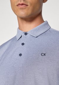 Svetlo modra polo majica z teksturirano tkanino, tremi temnimi gumbi in vezenim logotipom "CK" na levem delu prsnega koša.