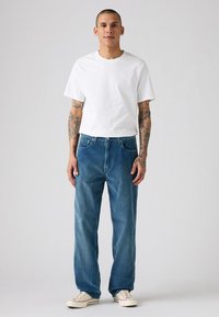 Un tricou alb cu mânecă scurtă, purtat împreună cu o pereche de blugi denim albaștri, având un croi lejer, o ușoară decolorare și un design cu cinci buzunare, completat cu pantofi off-white.