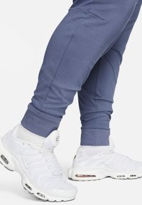 Witte Nike sneakers met luchtkussens, gecombineerd met strakke blauwe joggingsbroeken. De broeken hebben elastische manchetten en zijzakken.