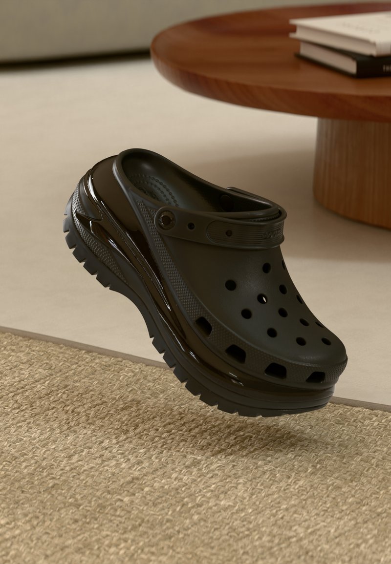 Chaussure noire de type sabot avec des trous de ventilation et une bride, flottant au-dessus d'un tapis beige texturé à côté d'une table ronde en bois.