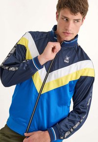 Blå, marinblå och gul löparjacka med dragkedja, chevrondesign och kontrasterande detaljer på ärmarna. Tillverkad av ett slätt syntetiskt material.