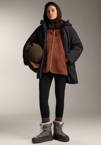 Black padded coat over a brown fleece top, black leggings, and gray insulated boots. Accessories include a brown knit scarf and a green bag.