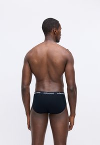 Muž s tmavou pokožkou otočený chrbtom, oblečený v čiernych Björn Borg šortkách na jednoduchom bielom pozadí.