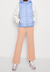 Jasnoniebieska kurtka puchowa z kapturem, pod spodem biała bluzka z długim rękawem, w połączeniu z brązowymi spodniami o prążkowanej fakturze i wielokolorowymi butami sportowymi.