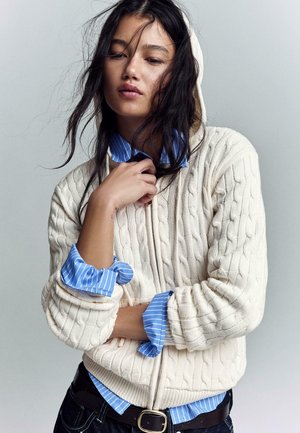 Mlada ženska z dolgimi temnimi lasmi nosi kremasto kapuco z vzorcem kitanja čez modro srajco s črtami, z eno roko prilagaja ovratnik, nevtralna siva podlaga.