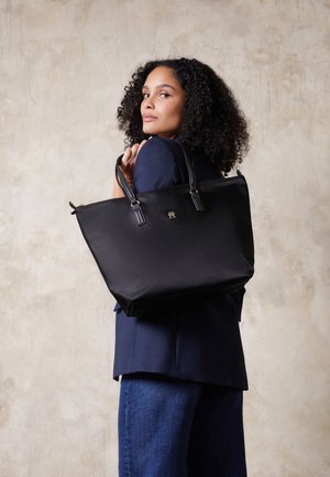 Frau mit lockigem Haar, die einen marineblauen Blazer und Jeans trägt und eine große schwarze Tragetasche über der Schulter vor einem einfarbigen beigen Hintergrund hält.