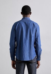 Pitkähihainen farkkupaita keskiharmaansinisessä sävyssä, napillinen kaulus, suora helma ja kalvosin yksityiskohdat. Kangas on sileää ja hieman kiiltävää.