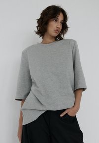 Szara, oversized koszulka z miękkiego, gładkiego materiału; ma krótkie rękawy, okrągły dekolt i lekko asymetryczny dół.