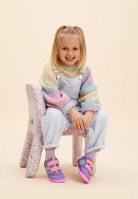 Pastellfarbener Pullover mit weiten Ärmeln, hellblaue Latzhose und lila Schuhe mit Klettverschluss, sitzend auf einem strukturierten Plastikstuhl.