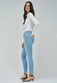 Jeans ajustados de color azul claro con cintura alta, bolsillos laterales y cierre con botón dorado. Combinados con una blusa blanca de manga larga y sandalias negras.