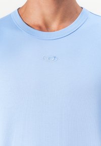 Close-up of a person wearing a light blue crew neck T-shirt featuring a small, subtly embroidered oval logo at chest center.