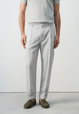 Homme portant un pantalon sur mesure gris clair avec revers, assorti à des mocassins en daim vert olive et une chemise à manches courtes gris clair.